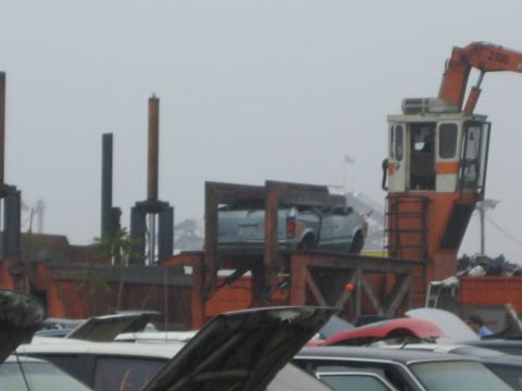 1990 Ford Bronco - orange crane marked 2100 hoists a gray pickup over rows of junked cars in a salvage yard under gray skies.