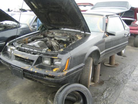 1990 Ford Bronco - weathered black Ford hatchback coupe sits on stands with its hood open and engine exposed in a wrecking yard.