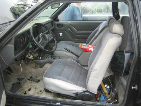 1990 Ford Bronco - gray cloth interior of an older coupe shows the steering wheel, bucket seats and a cluttered floor through the open driver door.