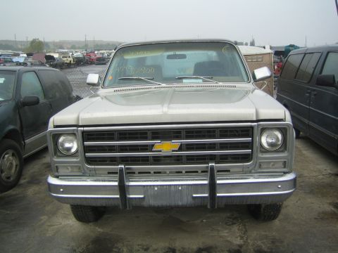 1990 Ford Bronco - tan square-body Chevrolet pickup with chrome grille and bumper faces forward in a foggy salvage lot.