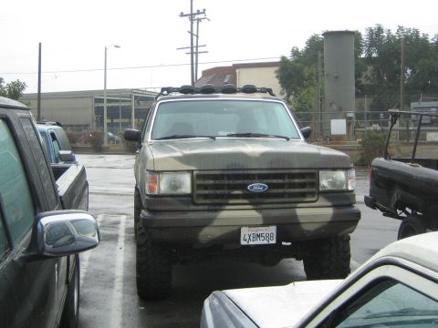 1990 Ford Bronco - tan lifted Ford SUV with a roof light bar and California plate 4XBM588 sits in a wet parking lot among other vehicles.