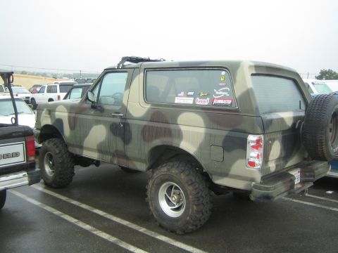 1990 Ford Bronco - camo-painted Ford Bronco with a spare tire and big mud tires sits parked on a gray day with decals on the rear glass.