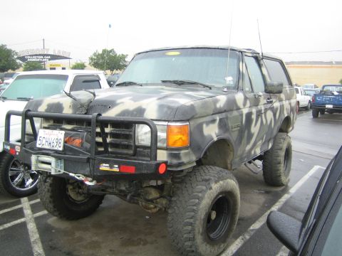 1990 Ford Bronco - camo-painted lifted Ford Bronco with brush guard, winch and California plate 5CHW698 sits in a lot near a Collector Car sign.
