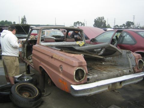 1990 Ford Bronco - man stands beside a rusty pink Ford Ranchero with its bed exposed and parts removed in a busy salvage yard.