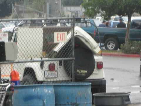 1990 Ford Bronco - white Hummer with its rear hatch open and a Las Vegas spare cover sits behind a chain-link fence and blue barrels.