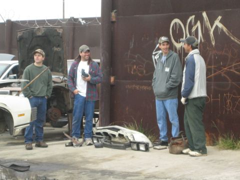 1990 Ford Bronco - Four men stand and talk beside a graffiti-marked rusty wall in a salvage yard with car parts on the ground.