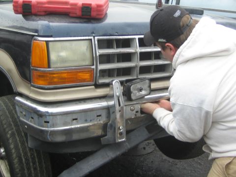 1990 Ford Bronco - man in a white hoodie and cap leans over the chrome front bumper and grille of a tan truck to remove parts.