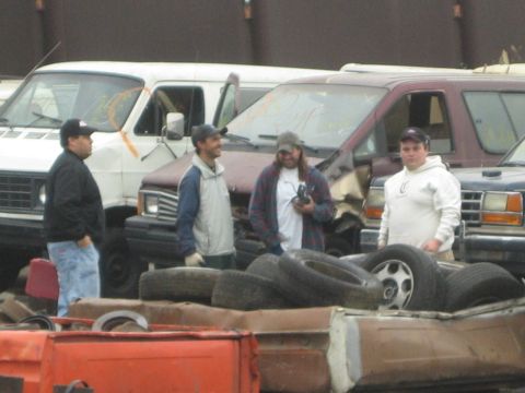 1990 Ford Bronco - Four men stand behind a pile of tires and wheels with white and maroon vans lined up behind them in a salvage lot.
