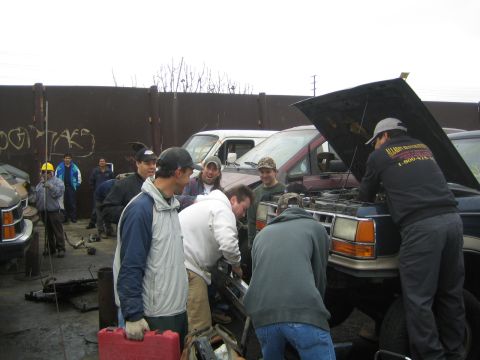 1990 Ford Bronco - group of men gathers around a maroon van with its hood up, working on the engine beside a graffiti wall in a salvage yard.