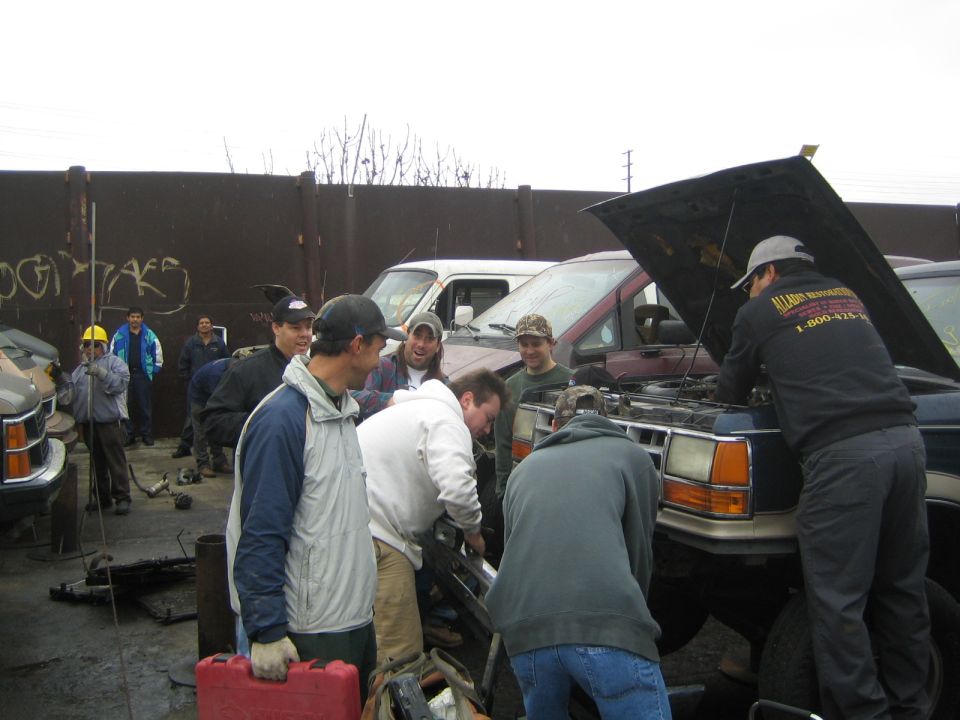 Group of men working on a maroon van with open hood in a salvage yard