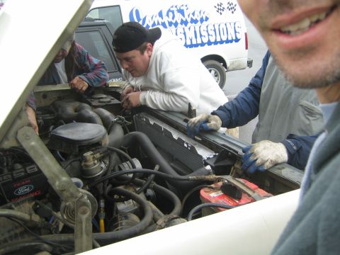 1990 Ford Bronco - Men in gloves work on an exposed engine and radiator under a raised white hood, with a Transmissions van in the background.