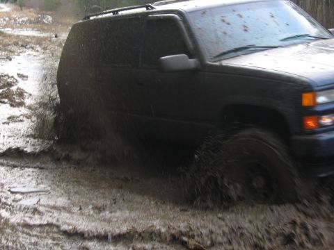 1976 GMC C/K Pickup - A dark SUV navigates a muddy terrain, kicking up water and mud as it drives through the mire.