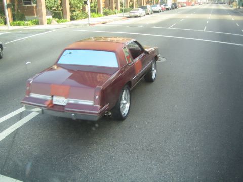 1990 Ford Bronco - maroon Oldsmobile Cutlass coupe on chrome wheels sits angled on a wide empty street lined with parked cars.