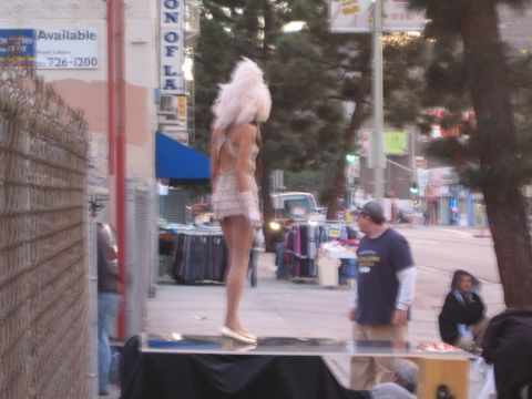 1990 Ford Bronco - street performer in a white feathered headdress and short sequined dress stands on a platform along a city sidewalk near vendors.