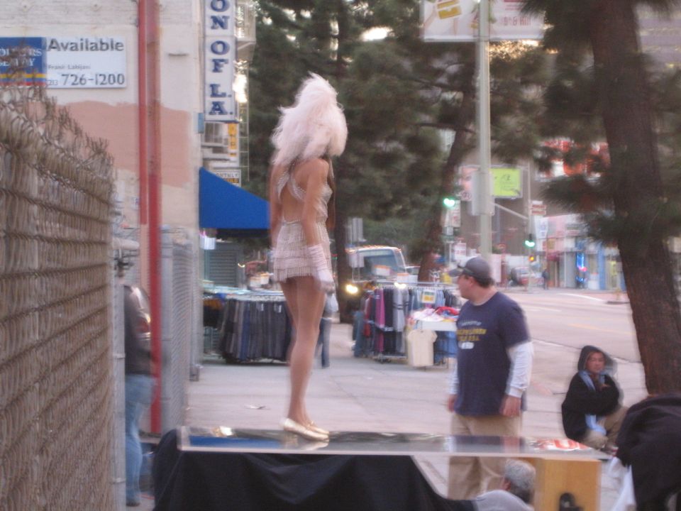 Performer in white feather headdress and sequined dress standing on platform on city sidewalk