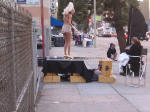 1990 Ford Bronco - performer in a sequined dress and blonde wig stands on a draped platform on a city sidewalk as people sit nearby.