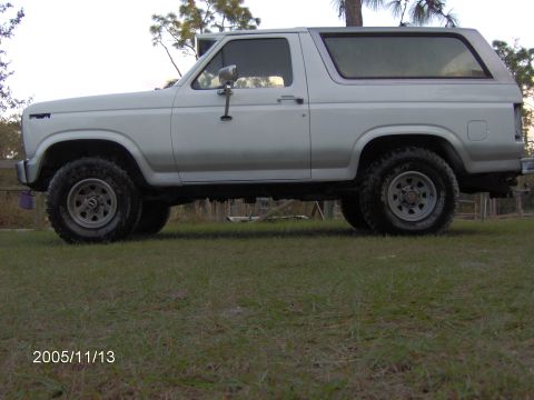 1983 Ford Bronco - A white SUV is shown from the side, parked on a grassy area with trees in the background.