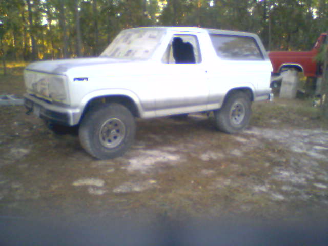 Silver lifted Ford Bronco SUV parked in a wooded dirt clearing, front driver-side view