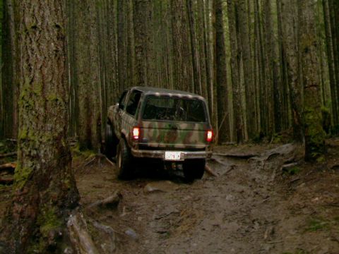 1987 Ford Bronco II - A camouflage SUV navigates a muddy trail through a dense forest.