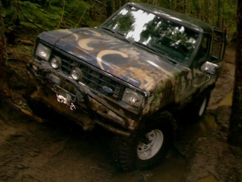 1987 Ford Bronco II - A 1987 Ford Bronco II navigates a muddy, wooded terrain with its door open.