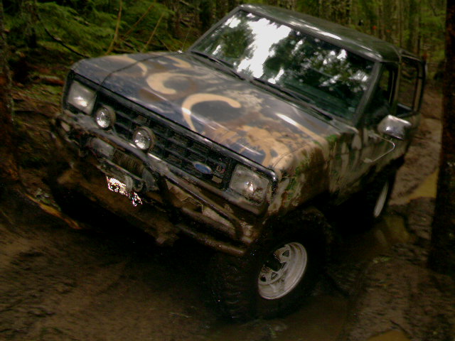 Muddy camo Ford 4x4 with winch and round driving lights in a forest rut