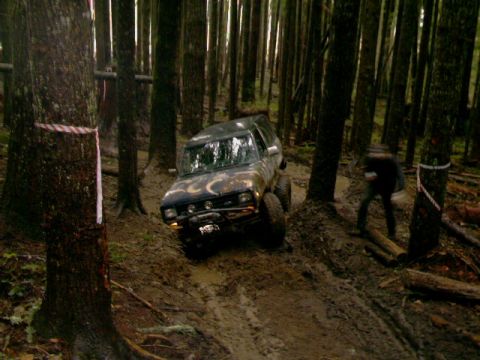 1987 Ford Bronco II - An off-road vehicle tackles a challenging muddy path through a dense forest, with a person visible in the background.
