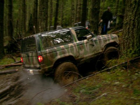 1987 Ford Bronco II - An off-road vehicle navigates a muddy trail in a dense forest environment.