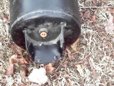1988 Ford Bronco - Close-up of a black cylindrical tank with a metal mounting bracket and an orange fitting, resting on the ground.