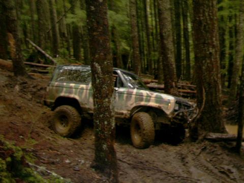1987 Ford Bronco II - This camouflaged SUV is navigating a muddy, wooded terrain with large tires.