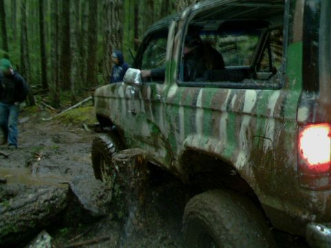 1987 Ford Bronco II - Mud-covered SUV navigates a difficult forest trail with visible damage.