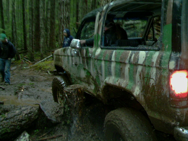 Camo lifted Bronco II driving through deep mud on a forest trail with onlookers watching