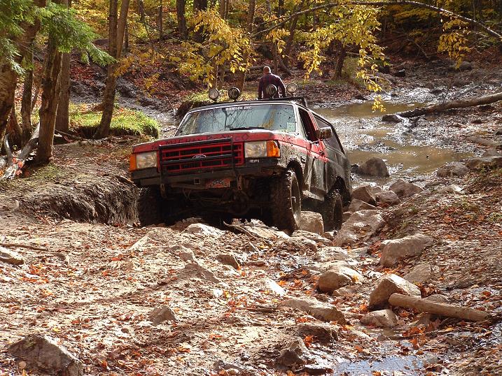 Red off-road SUV with roof rack driving through a rocky stream in a forest