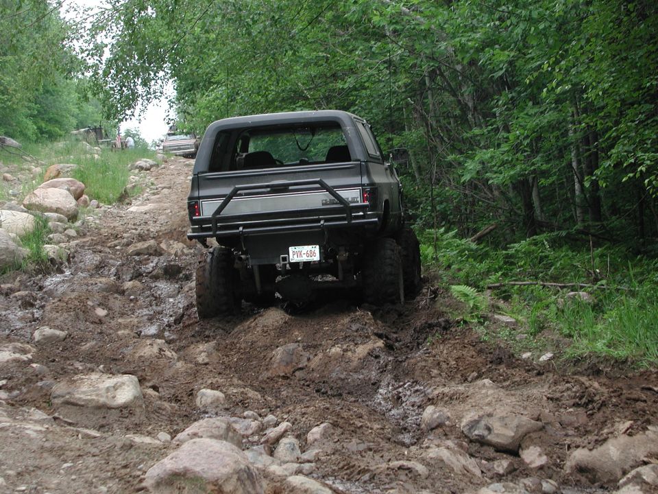 Dark SUV with a roll cage going up a muddy, rocky trail. Another SUV is visible ahead