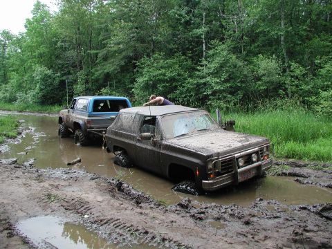 1977 Ford F-150 - A dark SUV is stuck in the mud while a blue pickup truck attempts to tow it out.