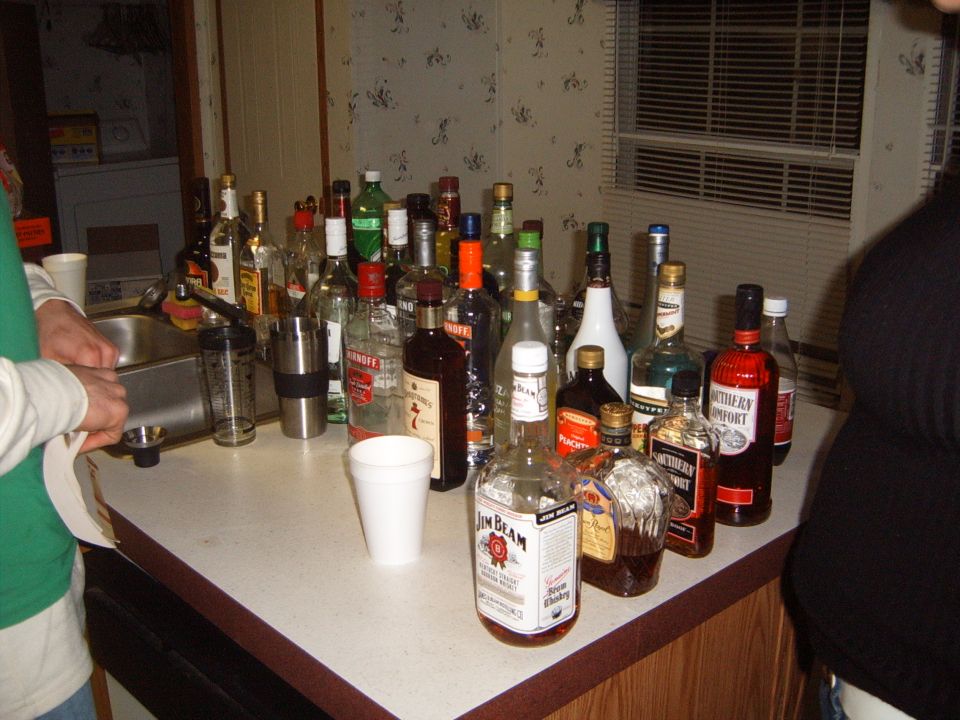 Lots of liquor bottles, including Jim Beam and Southern Comfort, on a counter with a sink and a person's hands visible