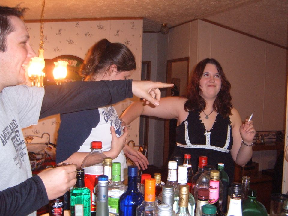 Three people are gathered around a bar with many bottles of alcohol. One person points to the right