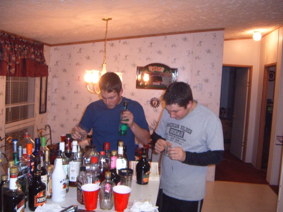 Two men pour drinks from bottles on a table laden with alcohol and red cups