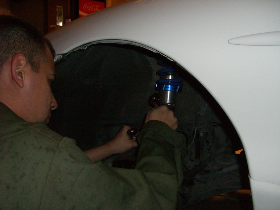 Man in green jacket adjusts blue coilover suspension on a white car's wheel well