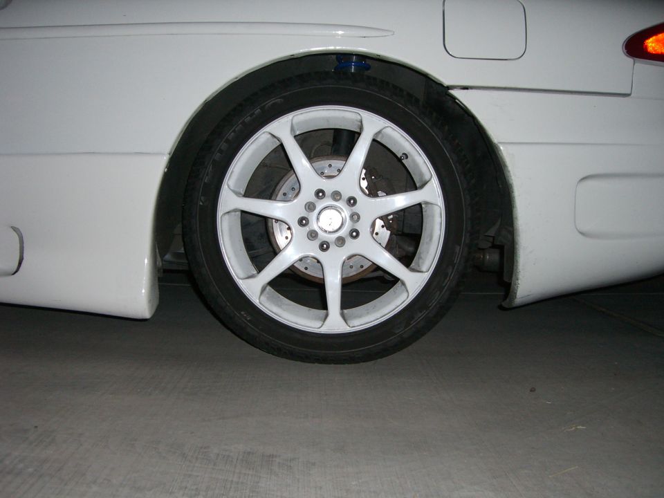 White car's rear wheel with a multi-spoke white rim and disc brake, showing the side fender and part of the bumper