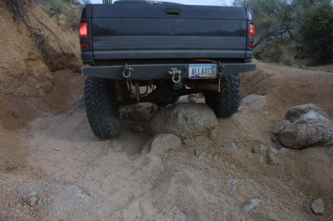 1994 Ford Bronco - This dark pickup truck is shown from the rear, tackling a challenging rocky and sandy terrain.