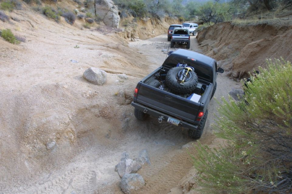 Several trucks and SUVs driving on a narrow dirt road in a rocky, arid landscape. The lead vehicle is a black pickup truck with a spare tire in the bed