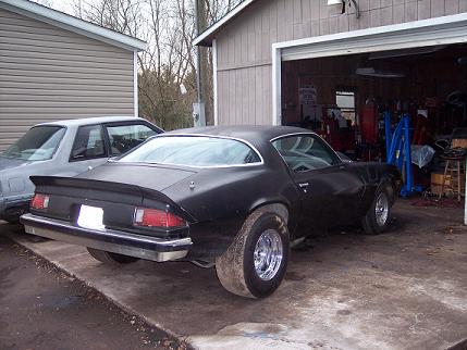 1977 Chevrolet Camaro - A black muscle car with wide rear tires is parked next to a garage, with another vehicle visible to its left.