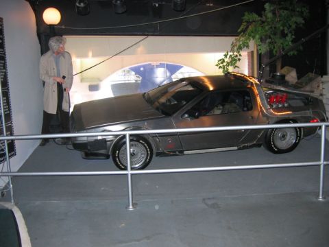 1985 Ford Crown Victoria LTD - A DeLorean DMC-12, recognizable by its unique gull-wing doors and stainless steel body, is displayed indoors.