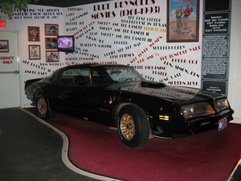 1985 Ford Crown Victoria LTD - A black Pontiac Trans Am is displayed indoors with walls covered in Burt Reynolds movie titles and memorabilia.