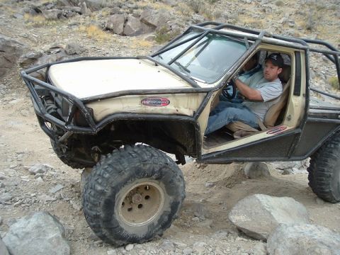 1989 Ford Bronco - A heavily modified off-road truck navigates a rocky trail with a driver at the wheel.