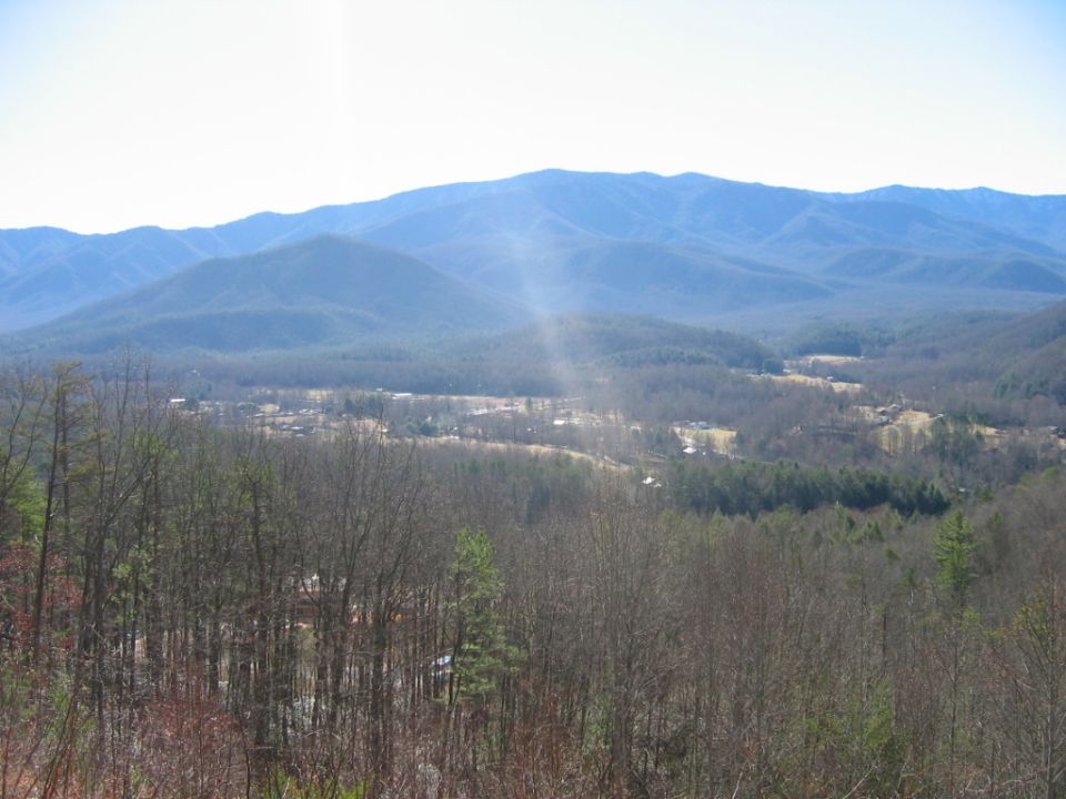 Mountainous landscape with bare trees in the foreground and a valley with buildings in the distance under a hazy sky