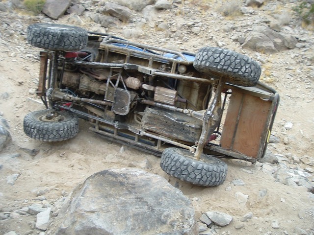 Rolled-over trail buggy on its side in a desert wash showing exposed frame and drivetrain