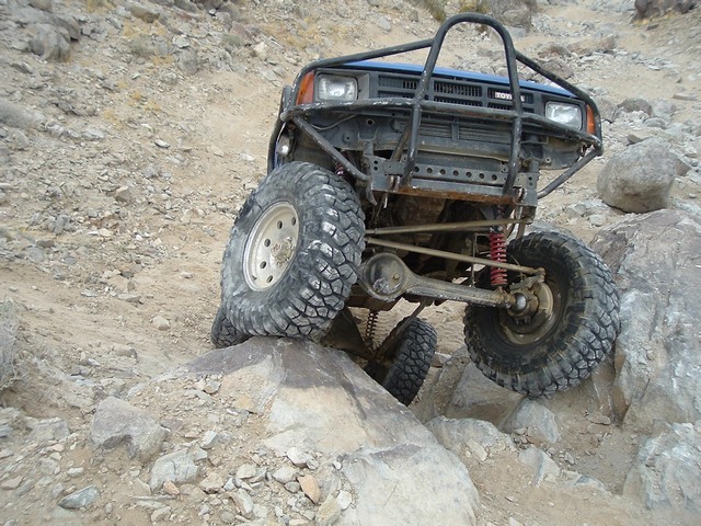 Blue Toyota truck with off-road tires climbing over rocks. It has a custom roll cage and bumper