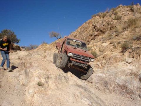 1989 Ford Bronco - A person watches a red Toyota truck ascend a challenging rocky terrain on a sunny day.