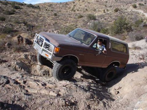 1989 Ford Bronco - A driver maneuvers a Ford Bronco over a challenging rocky trail in a desert landscape.